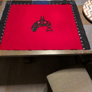 May include: A red textile with a black border and a central design of a black orca whale. The border is adorned with silver studs. The textile is displayed on a wooden table.