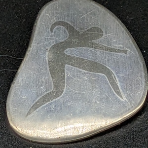 May include: A silver-coloured, pebble-shaped pin featuring a stylised figure of a person in motion. The figure is etched into the surface, creating a dark silhouette against the lighter background. The pin has a smooth, rounded edge.