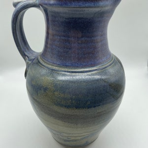 May include: A ceramic pitcher featuring a rounded body and a curved handle. The upper portion is glazed in a deep, vibrant blue, while the lower section transitions into a muted, earthy grey-blue tone with subtle horizontal texture and variations.