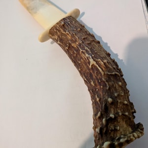 Puede incluir: Un cuchillo de caza con hoja y mango. La hoja es blanquecina y el mango es de un material marrón texturizado, posiblemente asta. El cuchillo tiene forma curva y una guarda.