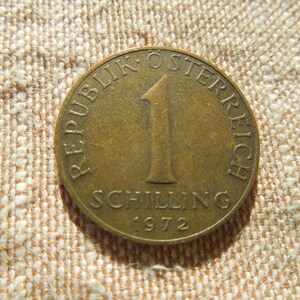 May include: A bronze Austrian 1 Schilling coin from 1972. The coin features the text "REPUBLIK OSTERREICH" and "SCHILLING" with the number "1" in the centre. The coin is set against a textured, beige background.