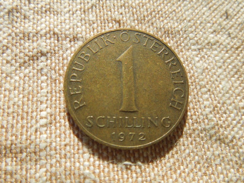 May include: A bronze Austrian 1 Schilling coin from 1972. The coin features the text "REPUBLIK OSTERREICH" and "SCHILLING" with the number "1" in the centre. The coin is set against a textured, beige background.