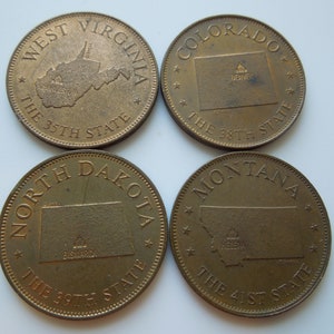 Puede incluir: Cuatro monedas conmemorativas circulares de tono bronce con mapas de estados de EE. UU. Las monedas están etiquetadas como West Virginia The 35th State, Colorado The 38th State, North Dakota The 39th State y Montana The 41st State.