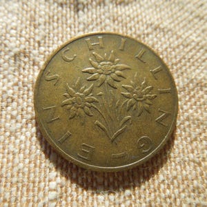 May include: A bronze coin with the word "SCHILLING" around the edge and three stylized flowers in the centre. The coin has a worn appearance and is set against a textured, light-coloured background.