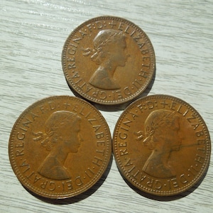 Puede incluir: Tres monedas de bronce con el perfil de una mujer y la inscripci&oacute;n "REGINA F.D. ELIZABETH II DEI GRATIA". Las monedas est&aacute;n dispuestas sobre una superficie de madera clara.