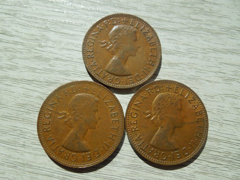 Puede incluir: Tres monedas de bronce con el perfil de una mujer y la inscripci&oacute;n "REGINA F.D. ELIZABETH II DEI GRATIA". Las monedas est&aacute;n dispuestas sobre una superficie de madera clara.