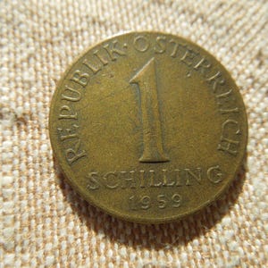 May include: A vintage Austrian 1 Schilling coin from 1959. The bronze coin features the text "REPUBLIK OSTERREICH" and "SCHILLING" around a large number "1". The coin is on a textured, beige surface.