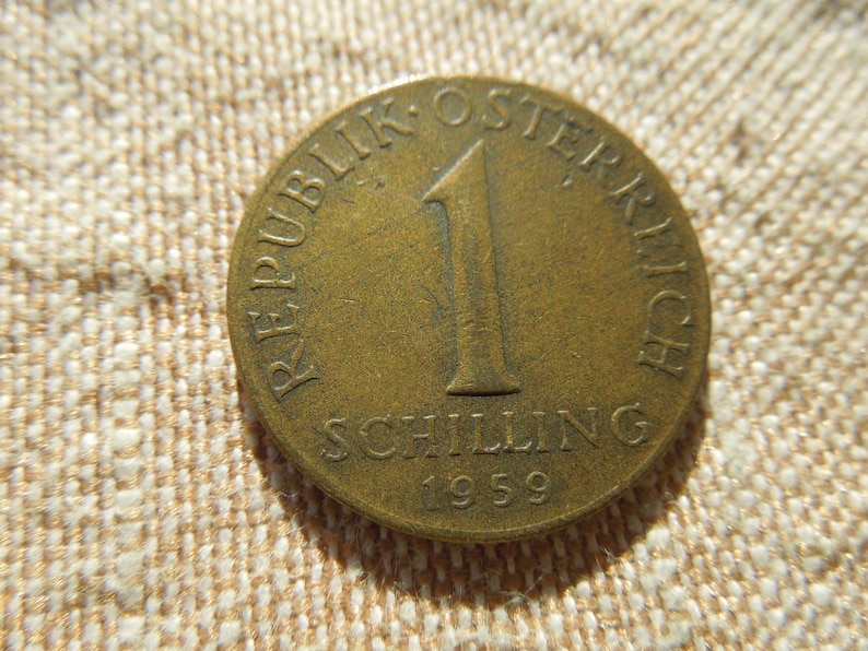 May include: A vintage Austrian 1 Schilling coin from 1959. The bronze coin features the text "REPUBLIK OSTERREICH" and "SCHILLING" around a large number "1". The coin is on a textured, beige surface.