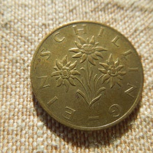 May include: A gold-coloured coin with three edelweiss flowers and the word "SCHILLING" around the edge. The coin is set against a textured, beige fabric background. The coin's design is detailed and well-preserved.