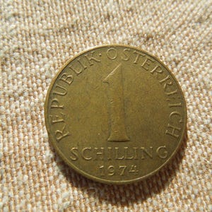 May include: A gold-coloured Austrian coin with the text "REPUBLIK OSTERREICH" around the edge. The coin features the number "1" and the word "SCHILLING" with the year "1974" at the bottom. The coin is on a beige fabric surface.
