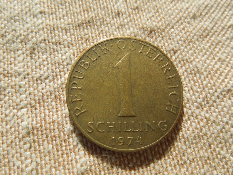 May include: A gold-coloured Austrian coin with the text "REPUBLIK OSTERREICH" around the edge. The coin features the number "1" and the word "SCHILLING" with the year "1974" at the bottom. The coin is on a beige fabric surface.