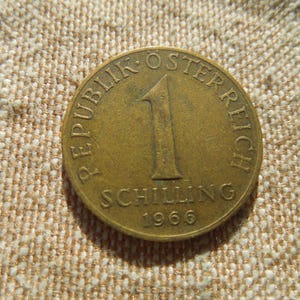 May include: A bronze Austrian 1 Schilling coin from 1966. The coin features the text "REPUBLIK OSTERREICH" and "SCHILLING" with the number "1" in the centre. The coin is on a textured, tan surface.