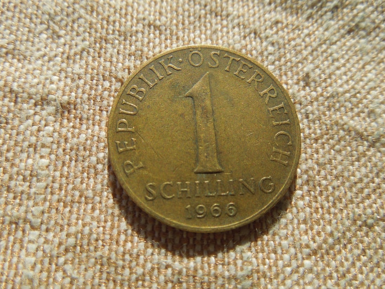 May include: A bronze Austrian 1 Schilling coin from 1966. The coin features the text "REPUBLIK OSTERREICH" and "SCHILLING" with the number "1" in the centre. The coin is on a textured, tan surface.