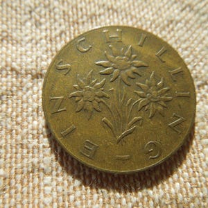 May include: A bronze coin with the word "SCHILLING" around the edge and a design of three stylized flowers. The coin is set against a textured, beige fabric background. The coin's design is embossed.