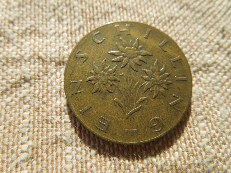 May include: A bronze coin with the word "SCHILLING" around the edge and a design of three stylized flowers. The coin is set against a textured, beige fabric background. The coin's design is embossed.