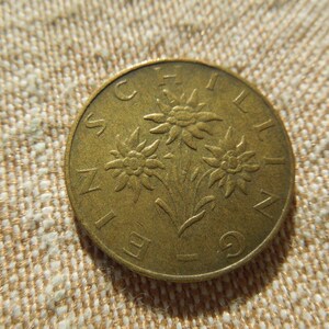 May include: A bronze coin with a design of three edelweiss flowers and the word "SCHILLING" around the edge. The coin is set against a textured, beige background. The coin's design is detailed and well-preserved.