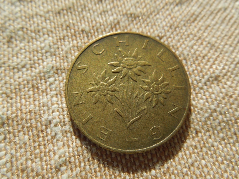 May include: A bronze coin with a design of three edelweiss flowers and the word "SCHILLING" around the edge. The coin is set against a textured, beige background. The coin's design is detailed and well-preserved.