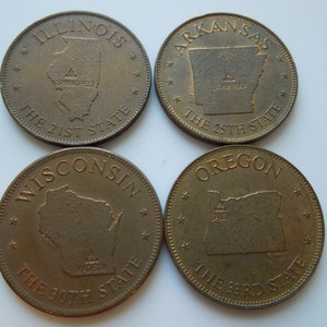 以下が含まれることがあります： イリノイ州、アーカンソー州、ウィスコンシン州、オレゴン州の州の輪郭を描いた、ブロンズ色のトークンが4つあります。各トークンには、州の名前、州都、州が連邦に加盟した順番が記載されています。