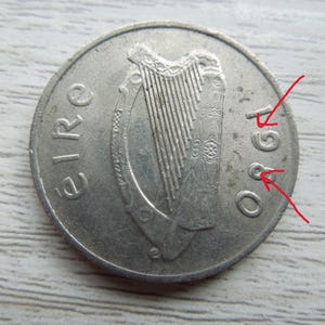 May include: A silver Irish coin from 1980, featuring a harp design. The word "eire" is inscribed on the coin's surface. The coin is circular and shows signs of wear.
