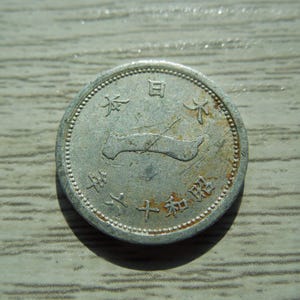 May include: A circular silver-toned Japanese coin featuring a central embossed map of Japan. The coin displays Japanese characters around the perimeter, including the text 日本 and 昭和六十六年, with visible signs of oxidation and surface wear.