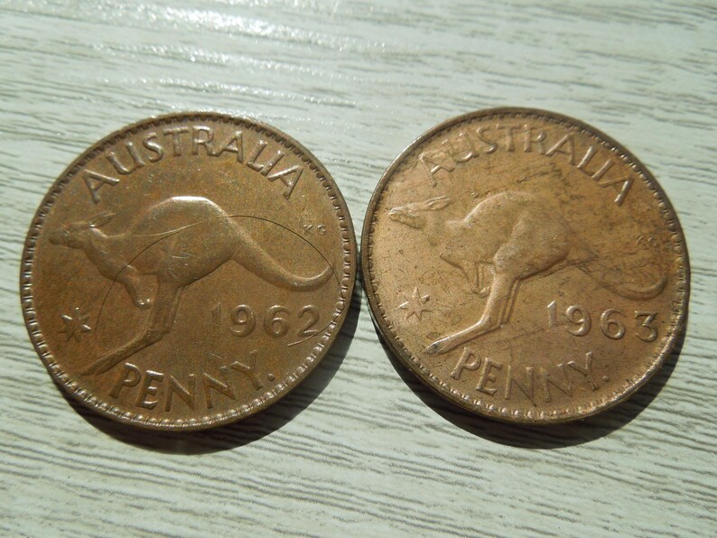 Puede incluir: Dos monedas australianas de un penique de color cobre. Presentan un canguro, las inscripciones &laquo; AUSTRALIA &raquo; y &laquo; PENNY &raquo;. Una moneda es de 1962, la otra de 1963. Las monedas est&aacute;n sobre una superficie gris claro.