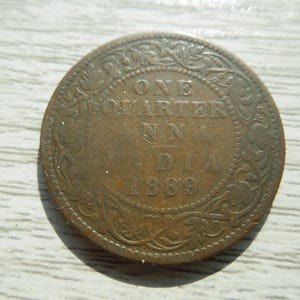 May include: A round, weathered copper-colored coin featuring the text ONE QUARTER ANNA INDIA 1889. The coin has a decorative floral border pattern encircling the central text, with a visible patina and signs of age on its metallic surface.