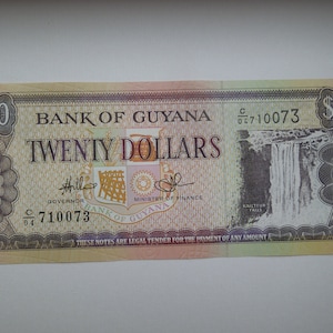 May include: A twenty-dollar banknote from the Bank of Guyana. The bill is predominantly tan with a waterfall image on the right. Text includes "Twenty Dollars", "Bank of Guyana", and "These notes are legal tender for the payment of any amount."