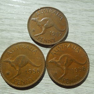 Puede incluir: Tres monedas antiguas de penique australianas. Cada moneda es de color cobre y presenta un canguro y la palabra "AUSTRALIA". Los a&ntilde;os 1957 y 1958 son visibles. Las monedas est&aacute;n sobre una superficie de veta de madera clara.
