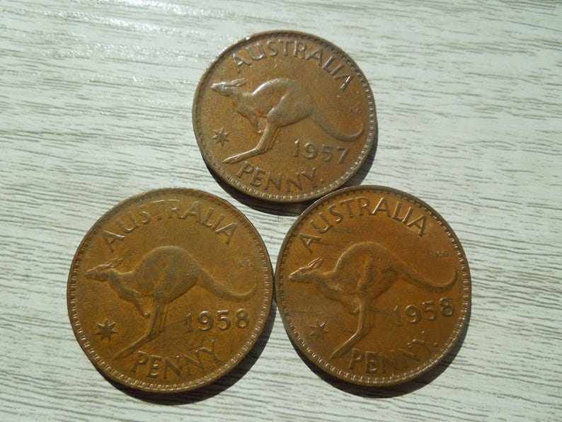 Puede incluir: Tres monedas antiguas de penique australianas. Cada moneda es de color cobre y presenta un canguro y la palabra "AUSTRALIA". Los a&ntilde;os 1957 y 1958 son visibles. Las monedas est&aacute;n sobre una superficie de veta de madera clara.