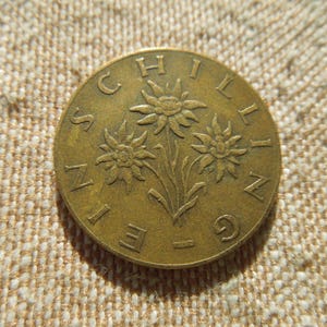 May include: A bronze coin with three edelweiss flowers and the word "SCHILLING" around the edge. The coin is set against a textured, beige background. The coin's design is in relief.