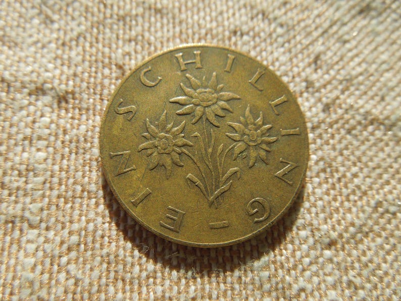 May include: A bronze coin with three edelweiss flowers and the word "SCHILLING" around the edge. The coin is set against a textured, beige background. The coin's design is in relief.