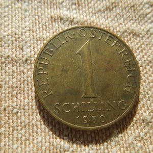 May include: A bronze Austrian 1 Schilling coin from 1980. The coin features the text "REPUBLIK OSTERREICH" and "SCHILLING" with the number "1" displayed. The coin is circular and has a slightly worn appearance.