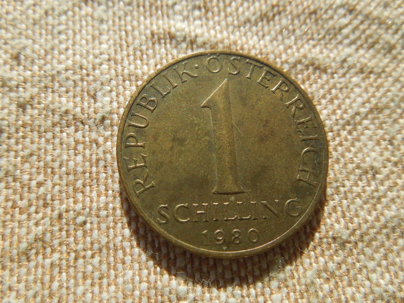 May include: A bronze Austrian 1 Schilling coin from 1980. The coin features the text "REPUBLIK OSTERREICH" and "SCHILLING" with the number "1" displayed. The coin is circular and has a slightly worn appearance.