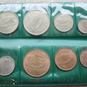 May include: A collection of Irish coins from the 1960s, displayed in a green plastic holder. The coins vary in size and color, with designs featuring a harp. The holder is sealed with clear plastic.