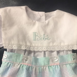 May include: A white baby onesie with a ruffled collar and a green embroidered "Bébé" on the front. The onesie is paired with a plaid diaper cover with pink, green, and blue squares.