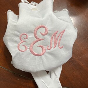 May include: White fabric bonnet with a pink embroidered "EM" monogram. The bonnet has a lace trim and white ties. The fabric appears to be soft and slightly gathered, with a button closure at the top.