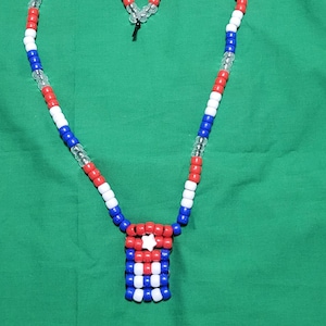 以下が含まれることがあります： 赤、白、青のビーズで作られたネックレスで、ハートの形をしています。ペンダントは白い星が付いた長方形のデザインです。ネックレスは緑色の背景にあります。