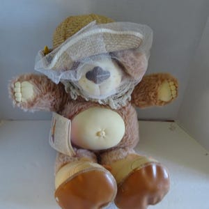 May include: A brown plush teddy bear wearing a brown straw hat with a white veil. The bear has a light brown nose and brown eyes. The bear is wearing brown boots.