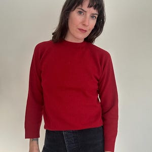 May include: A solid red sweater with long sleeves. The sweater has a crew neck and is made of a soft material. The person in the image is wearing black pants. The background is a plain white wall.