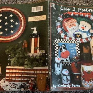 May include: A book titled "Luv 2 Paint for the Seasons" by Kimberly Parks. The cover features a painted design of a snowman and Santa Claus in a red and white candy cane wreath. The book also includes a painted design of a cow with the text "Cow lovers are like..."