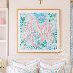 May include: A square wall art print in a gold frame featuring a large pink monogram letter design centered over a background of blue and green floral and botanical illustrations. The artwork is displayed above a bed with white and blue patterned pillows.