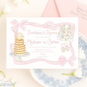 Op de afbeelding: Een lichtroze uitnodiging met de tekst "Pancakes & Pajamas" en "Madison is Seven". De uitnodiging bevat illustraties van pannenkoeken, pyjama's en strikken. Een roze envelop en een bloemenbord zijn ook zichtbaar.