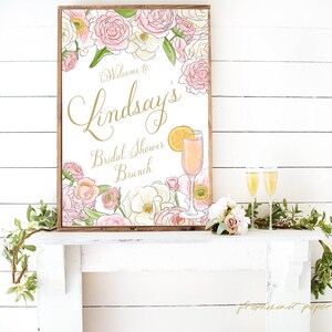 May include: A white sign with a gold frame and floral border. The sign says "Welcome to Lindsay's Bridal Shower Brunch". A mimosa is pictured next to the text.