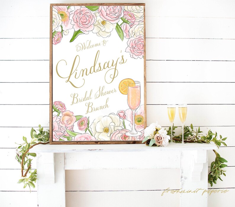 May include: A white sign with a gold frame and floral border. The sign says "Welcome to Lindsay's Bridal Shower Brunch". A mimosa is pictured next to the text.