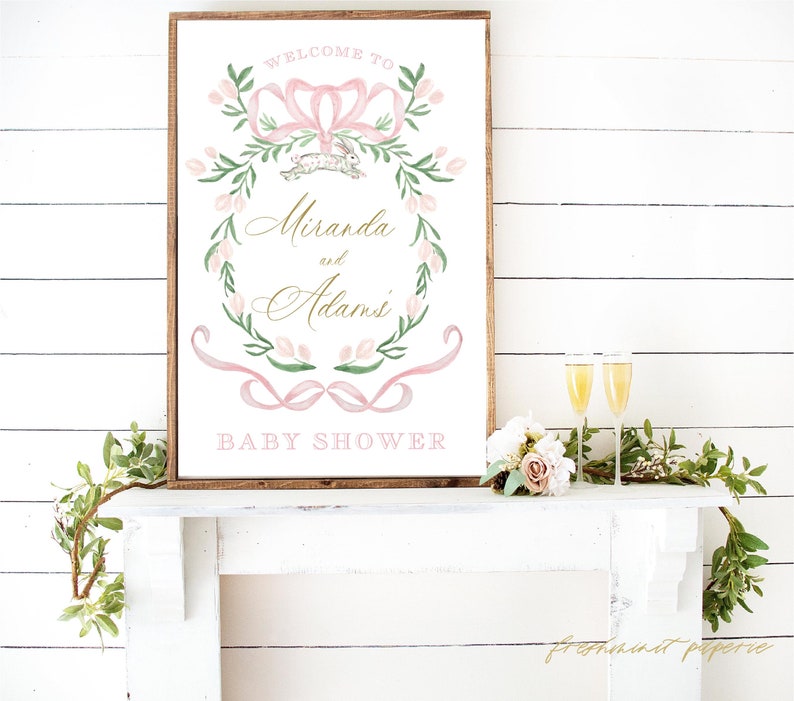 Op de afbeelding: Een roze en groene bloemenkrans met een roze strik en een wit konijn. De krans is op een witte achtergrond met de tekst "Welkom bij Miranda en Adams Baby Shower".