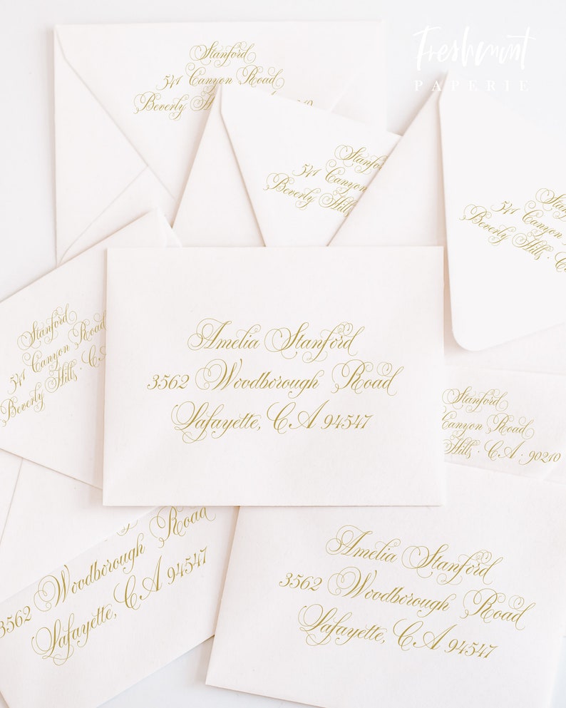 May include: A collection of ivory envelopes with gold calligraphy. The envelopes feature names, addresses, and the text "Freshmint Paperie." The calligraphy is in a flowing script, perfect for wedding invitations or announcements.