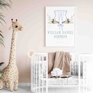 May include: A white crib with a brown blanket and white pillows. A stuffed giraffe stands next to the crib. A framed print with a blue and white monogram and the name "William Daniel Johnson" is on the wall.