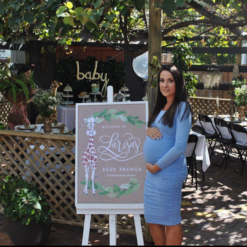 May include: A baby shower scene with a sign that reads "Welcome to Larisa's Bebe Shower." The sign features a giraffe illustration and is on an easel. A woman in a light blue dress stands next to the sign, holding her baby bump. The background includes decorations and a table setting.