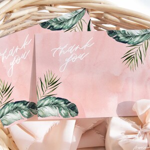 May include: Two pink thank you cards with green tropical leaf designs and white calligraphy. The cards are in a wicker basket with pink fabric.