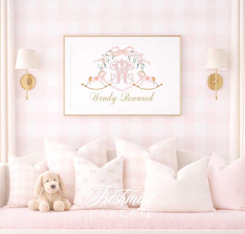 May include: A framed print with a gold border featuring a pink and gold design. The design includes a floral wreath, rocking horses, and the name "Wendy Rosewood" in gold script. The print hangs above a pink couch with pillows.